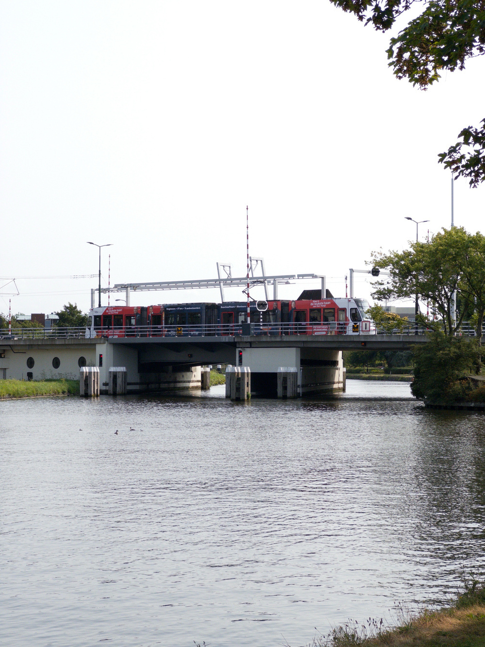 Auto-generated description: A red train is crossing a bridge over a body of water, with trees and streetlights visible in the background.