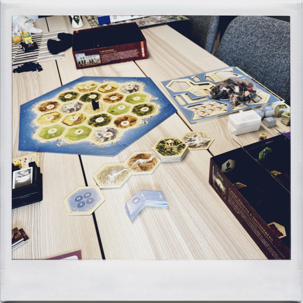 Auto-generated description: A tabletop game setup features a hexagonal board with various terrain types, cards, tokens, and game boxes arranged on a wooden table.