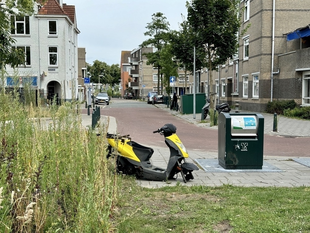 A yellow and black scooter without wheels is parked on a sidewalk near a residential street with buildings and greenery in the background.