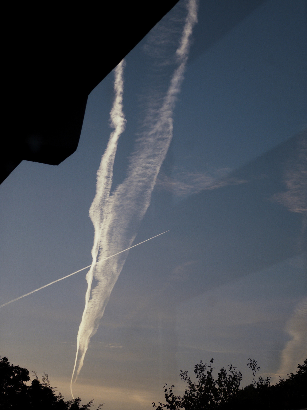 Visible in the sky are several distinct contrails from aircraft, set against a backdrop of a partially illuminated blue sky with silhouetted trees at the bottom.