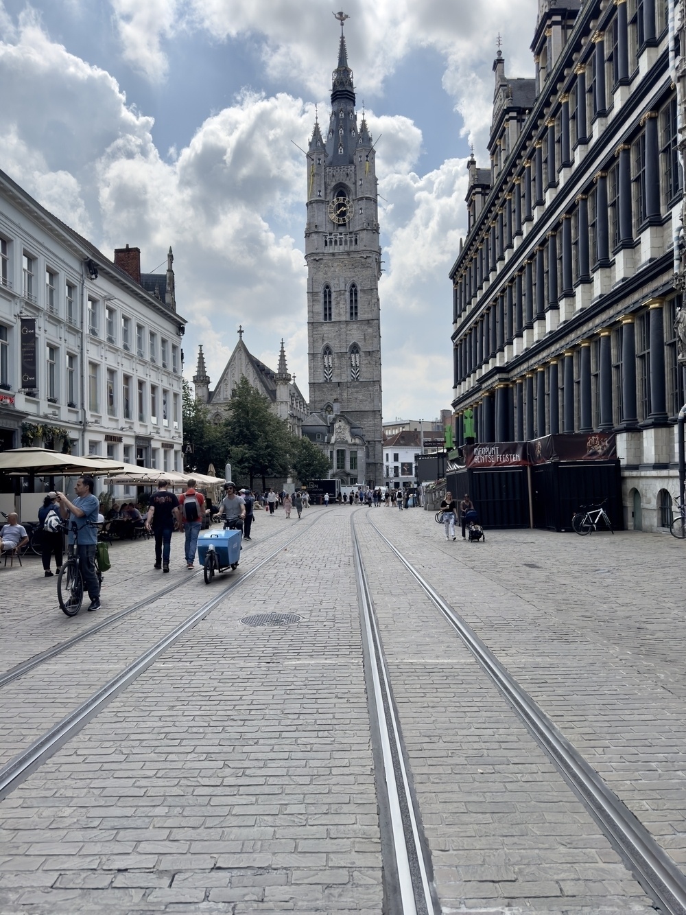 A cobblestone street with tram tracks runs between historic buildings, leading to a tall clock tower in the distance, while people walk, bike, and socialize alongside.