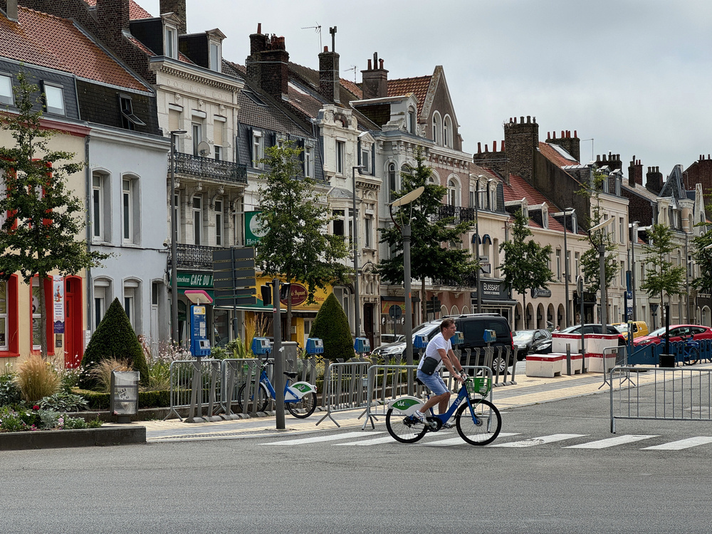 A street scene in Calais, France, featuring a row of charming, historic buildings with varied architectural styles and colorful facades. In the foreground, a man in casual attire rides a blue bicycle across a pedestrian crossing. The street is lined with trees, parked cars, and a few barriers. The atmosphere is calm and picturesque.