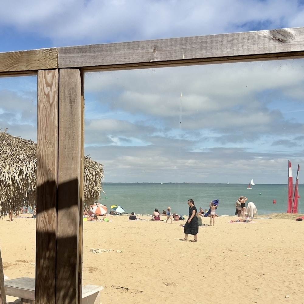 A sunny beach scene with several people relaxing on the sand, a sailboat in the distance, and a thatched hut structure partially visible in the foreground.