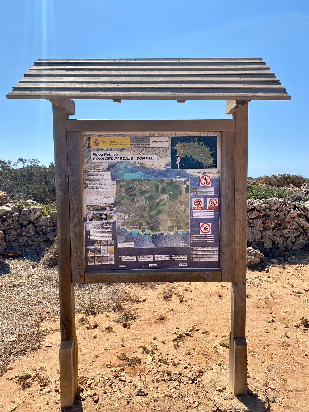 Wooden informational signboard at Cova des Pardals - Son Vell in a natural, rocky landscape. The sign includes a map, text details about the area, regulations, and symbols indicating prohibited activities. The sky is clear and blue.