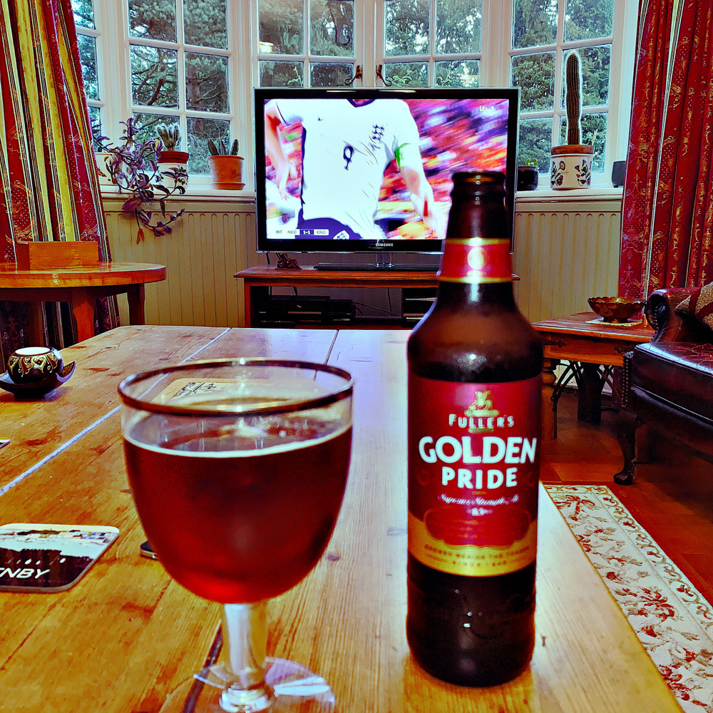 A cozy room with a wooden table, on which sits a glass of dark beer and a bottle of Fuller's Golden Pride. In the background, a television is showing a soccer match with a player in a white jersey with the number 9 on the chest