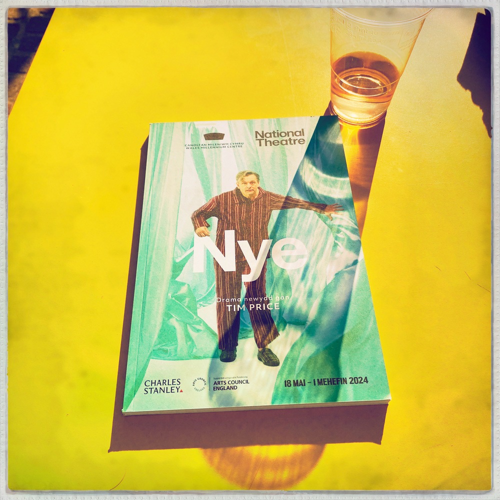 A yellow table with a theatre programme from The National Theatre titled Nye placed on it. The programme mentions a new drama by Tim Price and displays a person in pyjamas posing dramatically.
