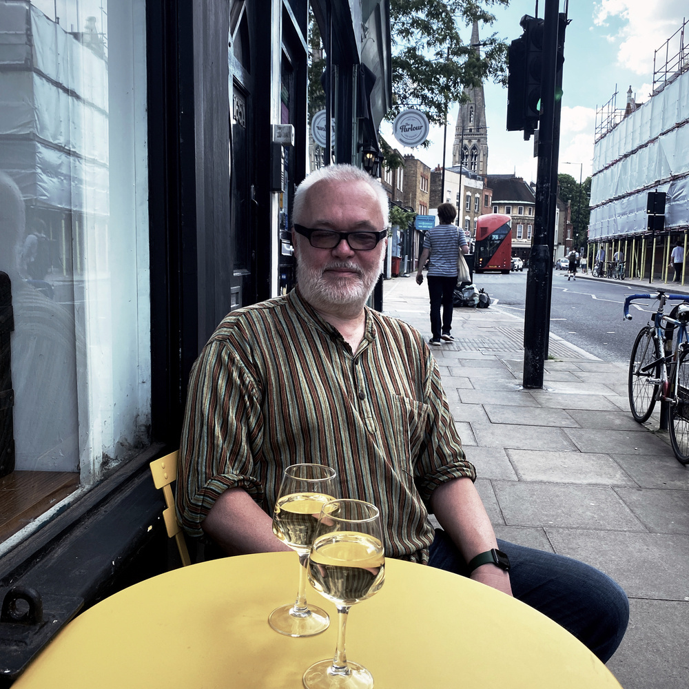 A man with white hair and a beard, wearing glasses and a striped shirt, sits at a round yellow table outside a café on a city street. Two glasses of white wine are on the table. In the background, there are buildings and a church.