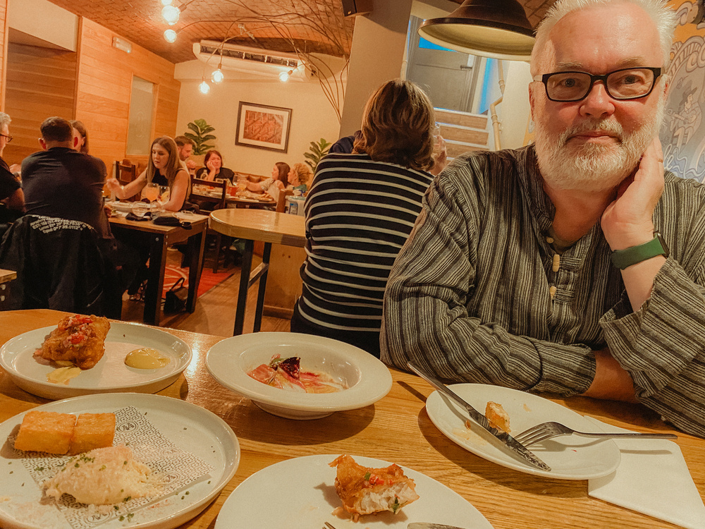 A bearded man is sitting at a restaurant table with several plates of food, looking towards the camera, while other diners are seated in the background.