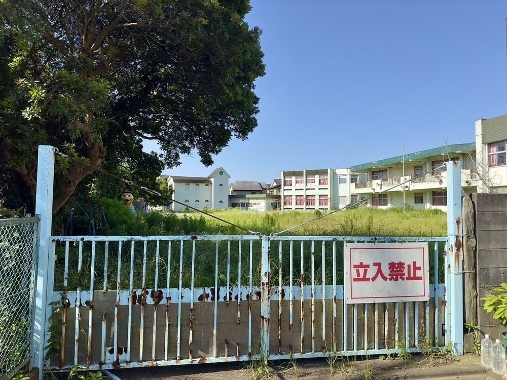 A rusty blue gate with a “no entry” sign in Japanese, stands in front of an overgrown grassy field and abandoned school buildings under a clear, sunny sky.