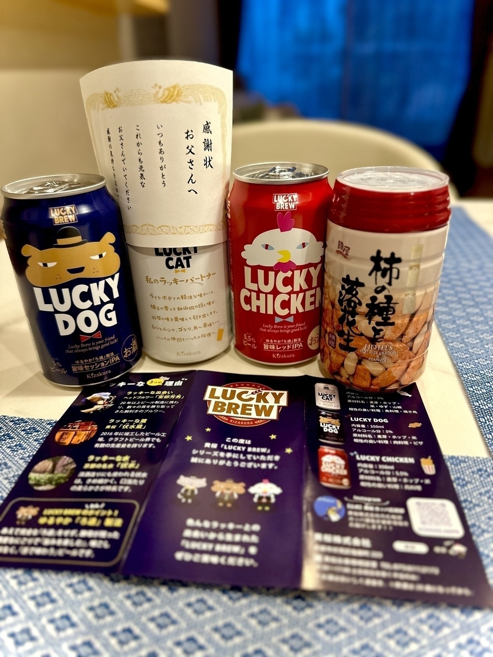 Several can beverages featuring "Lucky Dog", "Lucky Cat", and "Lucky Chicken" designs are displayed, with informational cards and a snack container in the background, all set on a blue-checkered tablecloth.