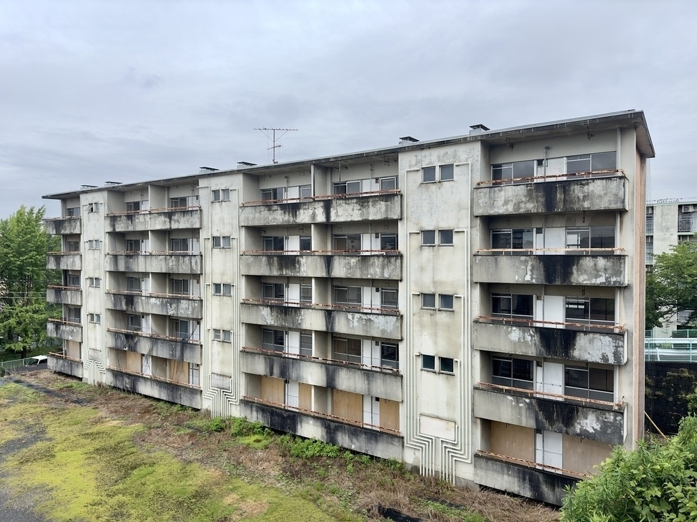 A dilapidated multi-story "danchi" public apartment building displays signs of heavy staining and neglect, with boarded-up windows and overgrown vegetation. In Yokohama Japan, under grey skies. 