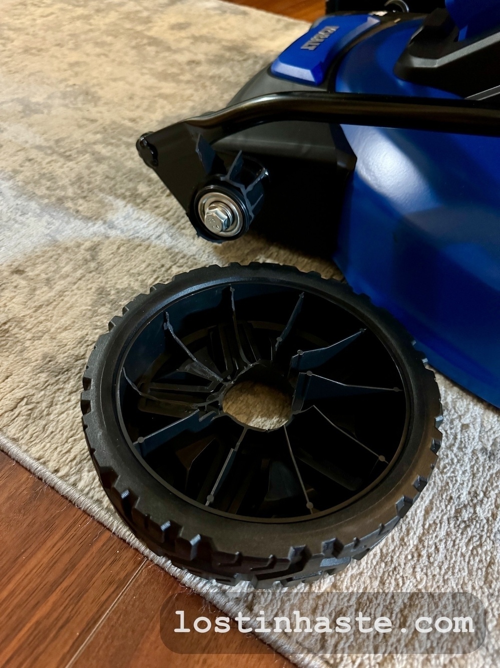 A detached black plastic wheel lies near the metal axle of a blue lawn mower, situated on a beige carpet next to a hardwood floor.