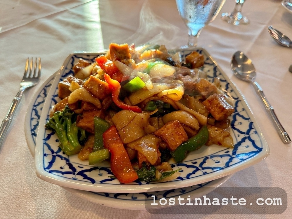 A steaming plate of stir-fried tofu and vegetables rests on a patterned dish, situated on a white tablecloth, accompanied by silverware. Text: 'lostinhaaste.com' is visible at the bottom right.