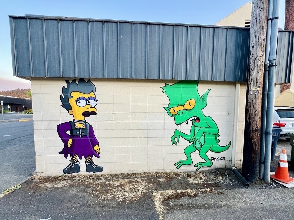 A wall with a mural of....characters from the Simpsons but I don't recognize them? On the left is some version of Goth Lisa in a purple dress with grey hair and fishnet sticking, and on the right is Bart but in some kind of green ghoul form. 