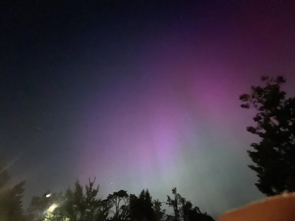 Pink and purple aurora in the night sky, over the roof of a house and the tops of some tree silhouettes. 