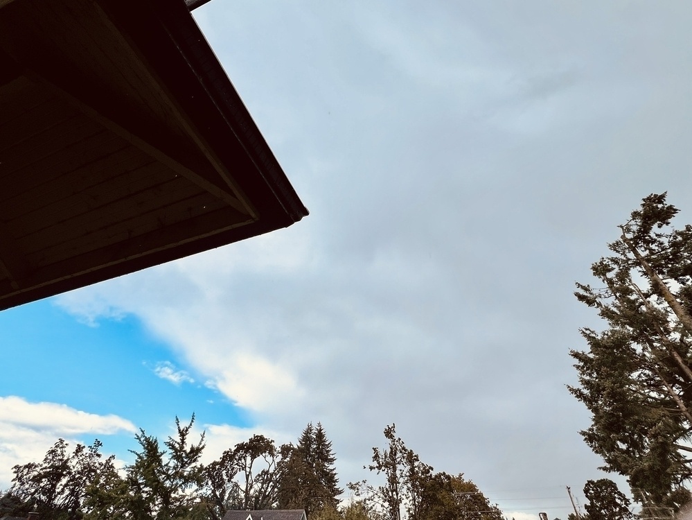 Big patch of sky, with a pointed dark corner of a roof laying over the upper left hand corner. The sky is fierce grey on the right but on the left is a beautiful bright blue patch peeking through the clouds.