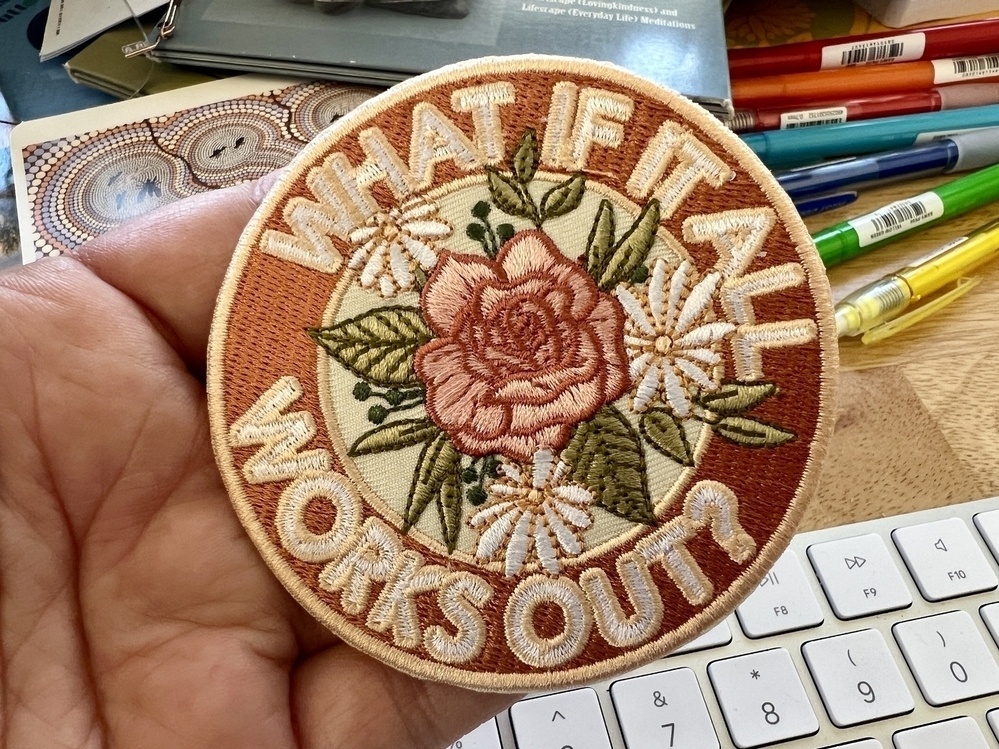 A round soft pink embroidered iron-on patch that says, WHAT IF IT ALL WORKS OUT? In the middle is a rose and three daisies.