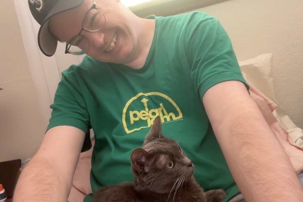 a white man in a baseball cap laughs while a grey cat looks on