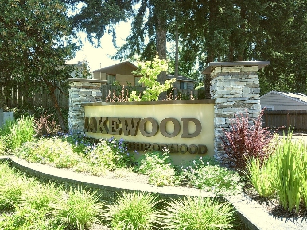 A sign that says Lakewood and Neighborhood (the other language is obscured) surrounded by a wall, columns, and a bunch of plants, on a sunny day 