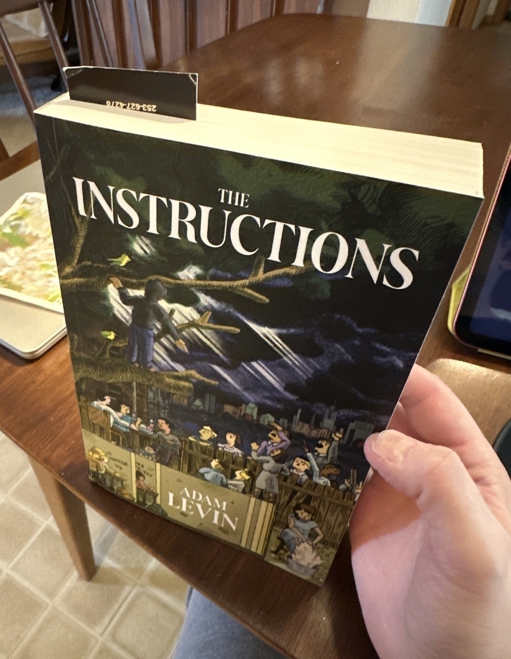 A man holds up the cover of the paperback book The Instructions, resting it on a wooden dining table