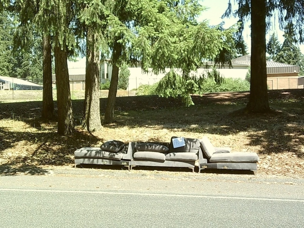 An outdoor scene with a beat up couch in the foreground that has a "free" sign on it 