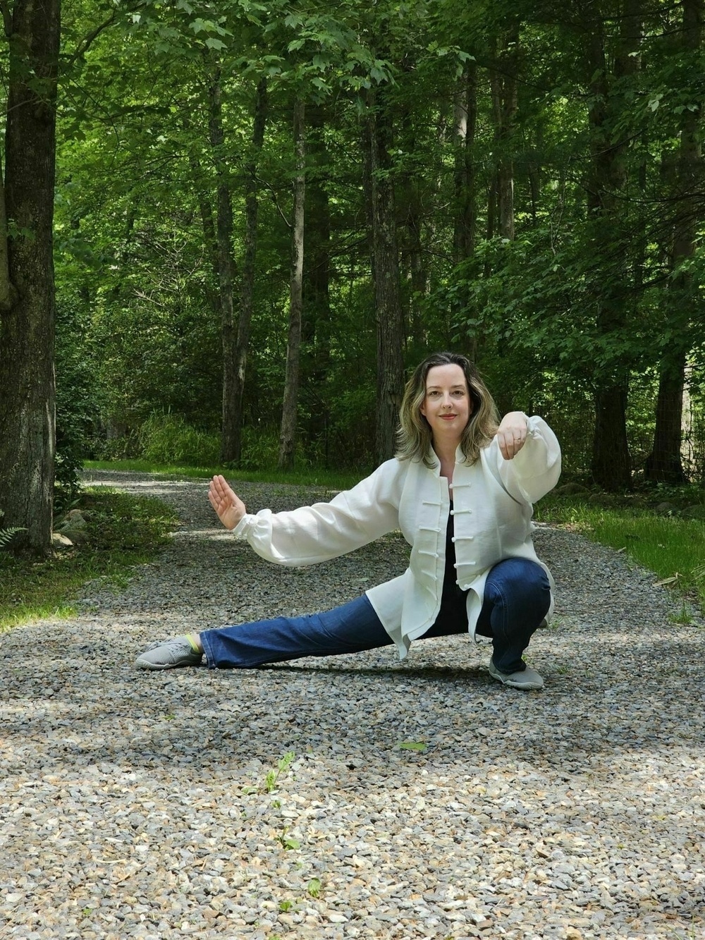 Auto-generated description: A woman performs a martial arts pose on a gravel path in a wooded area.