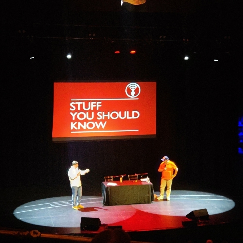 Auto-generated description: A stage setup features a large screen displaying the text “Stuff You Should Know,” with two individuals standing beside a table in the spotlight.