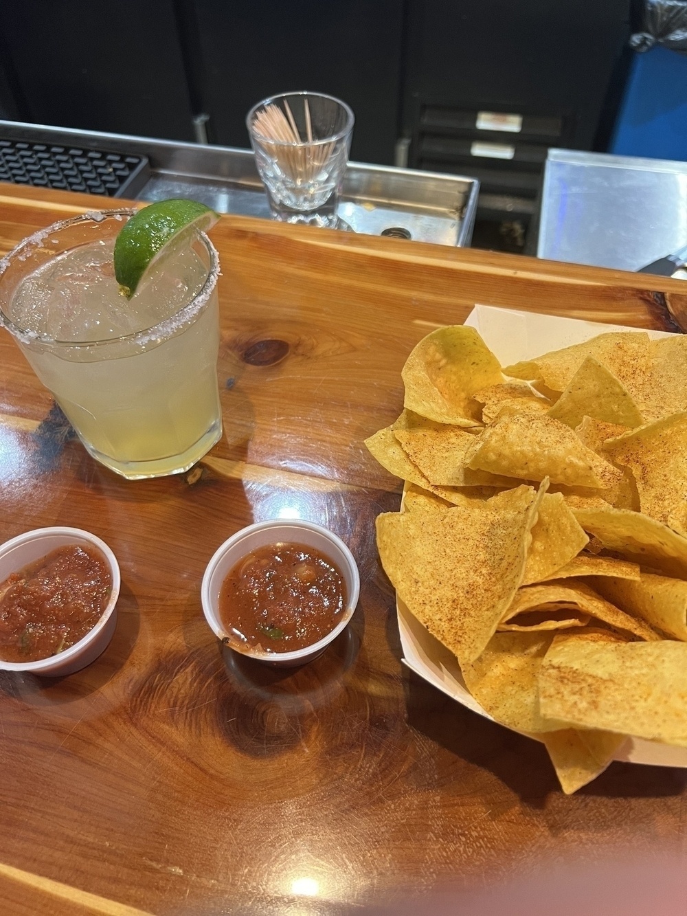 A margarita in a rock class with a salted rim and a lime side beside an order of chips and a few little containers of salsa on a wooden bar.