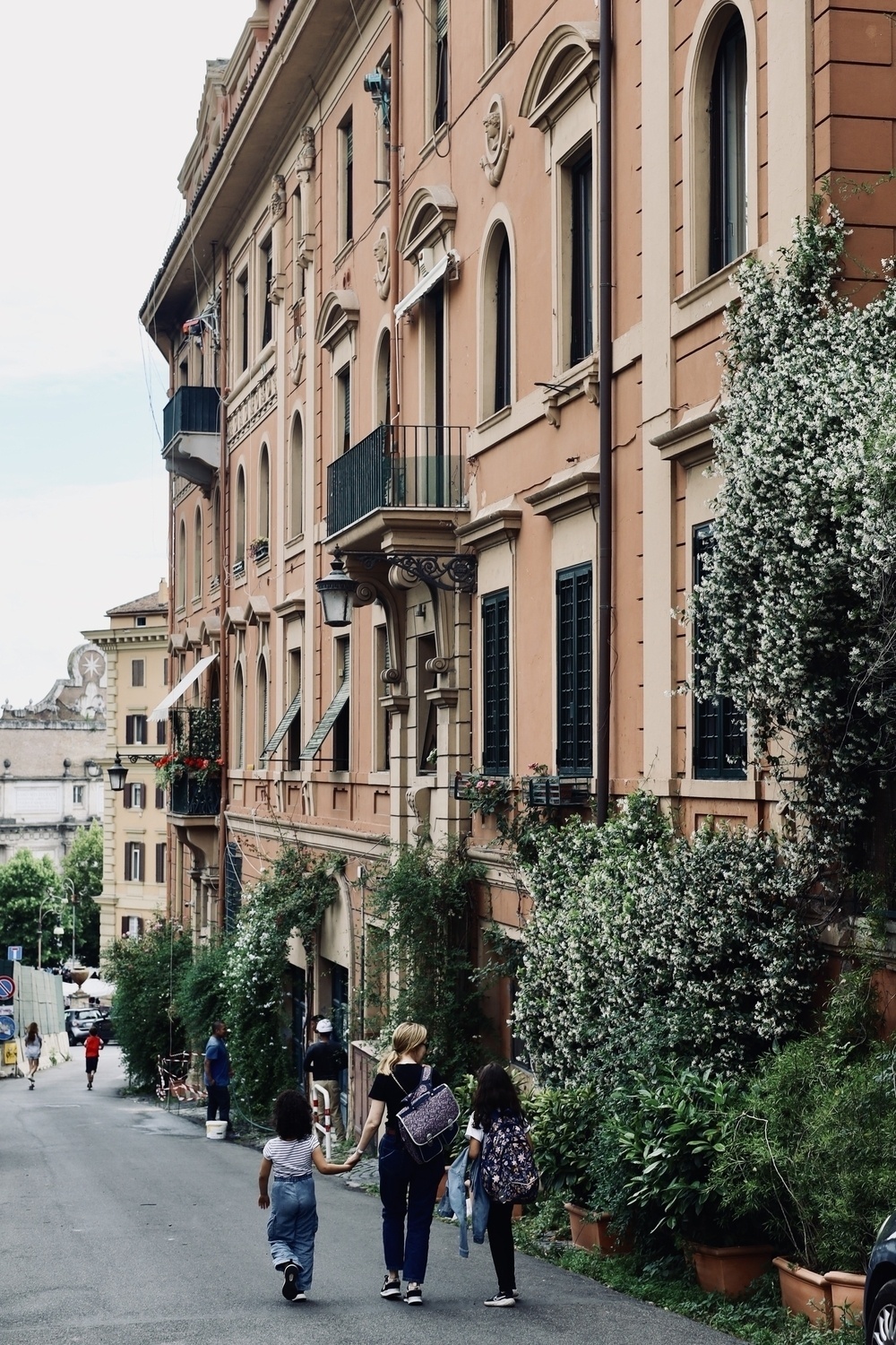 A group of people strolls down a quaint, plant-lined street flanked by charming, historic buildings.