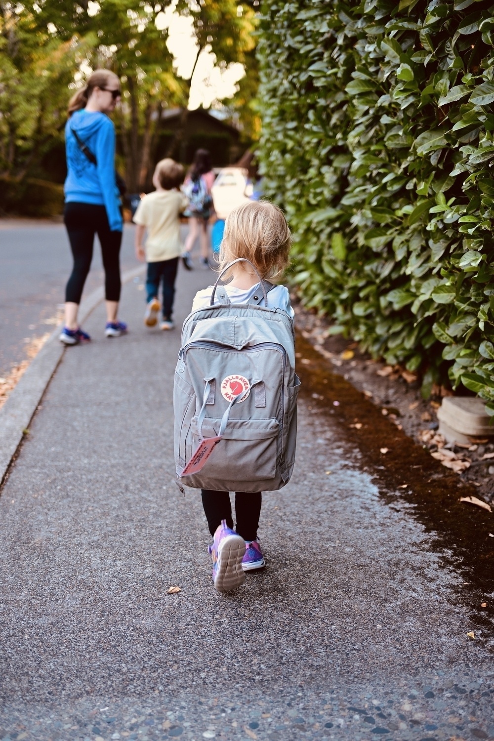 A young child with a large backpack walks along a sidewalk, followed by a woman and another child.