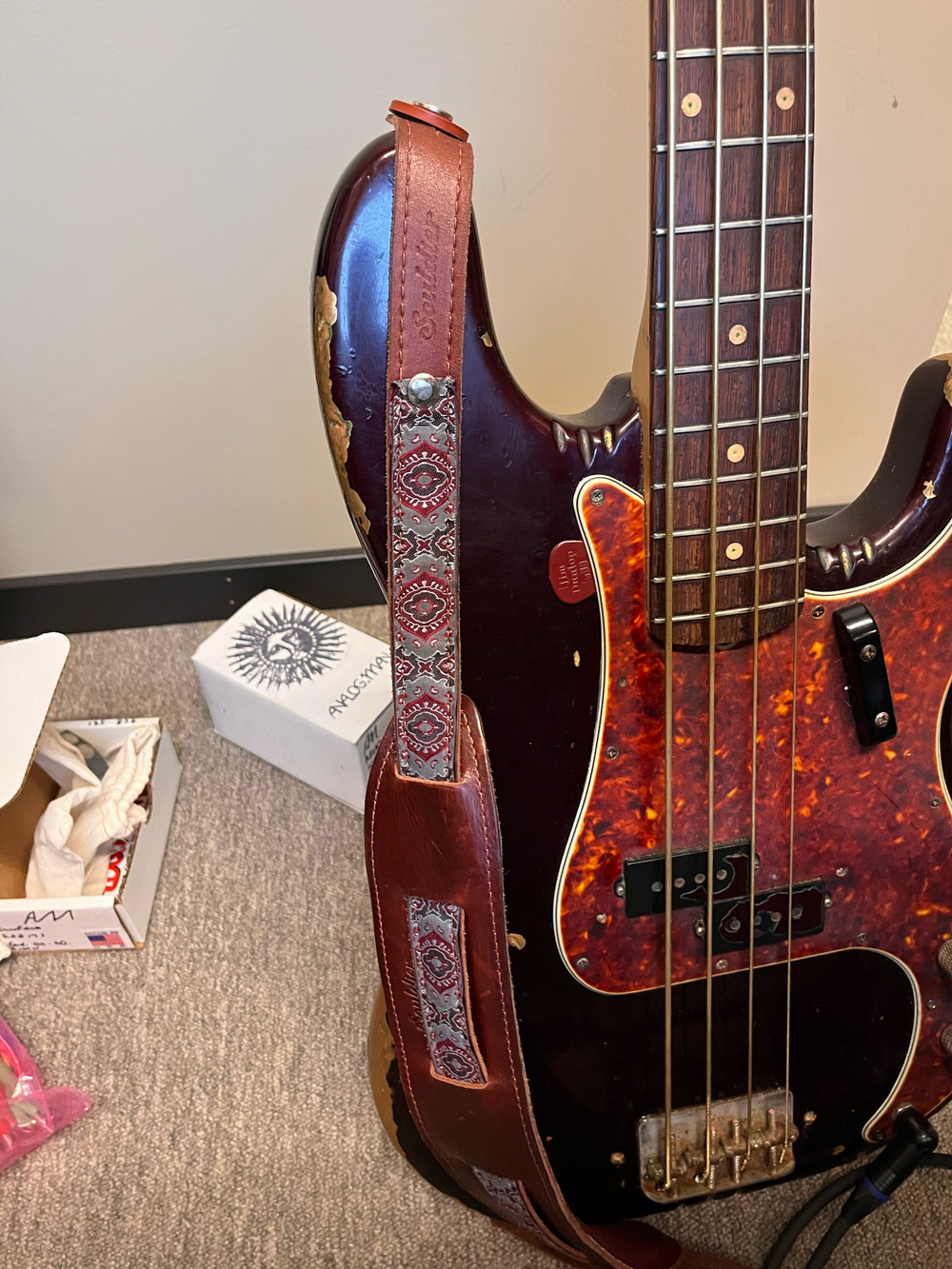 Fender P-bass from 1961, with a burgundy leather strap from Souldier. The bass is a very dark purple with a tortoise pick guard. Someone has made notches in some places.