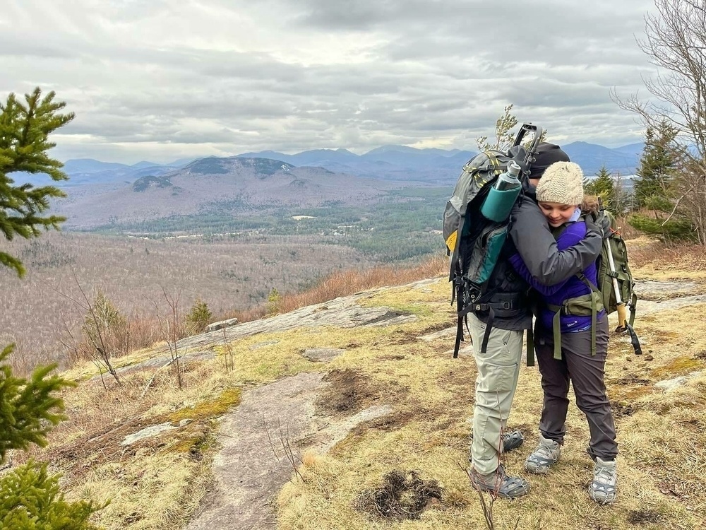 Two hikers are embracing on a mountain trail, with a scenic overlook of forested hills and cloudy skies in the background. There is no text in the image.