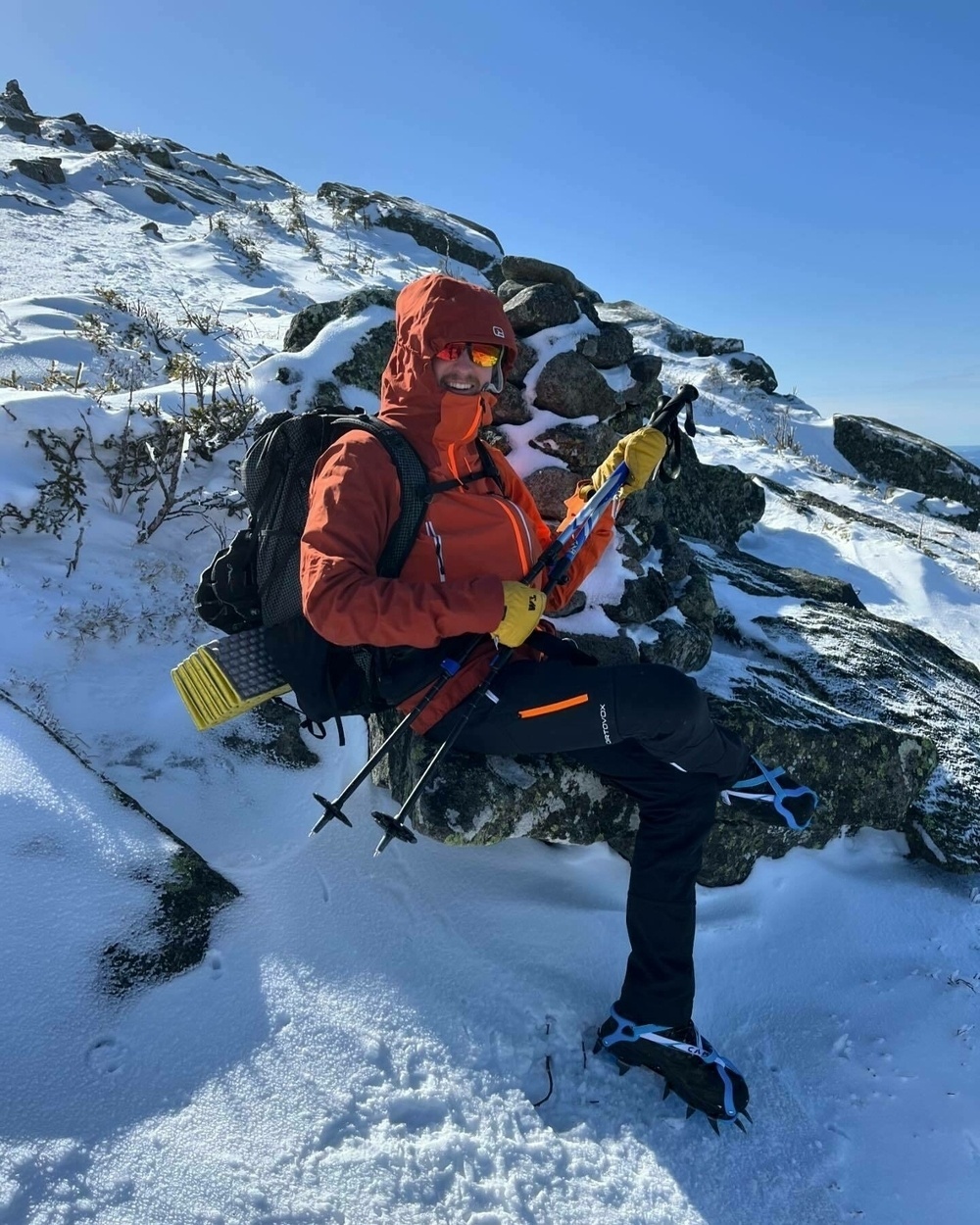 A person in winter clothing and gear is resting on a snowy mountain slope with clear blue skies in the background.