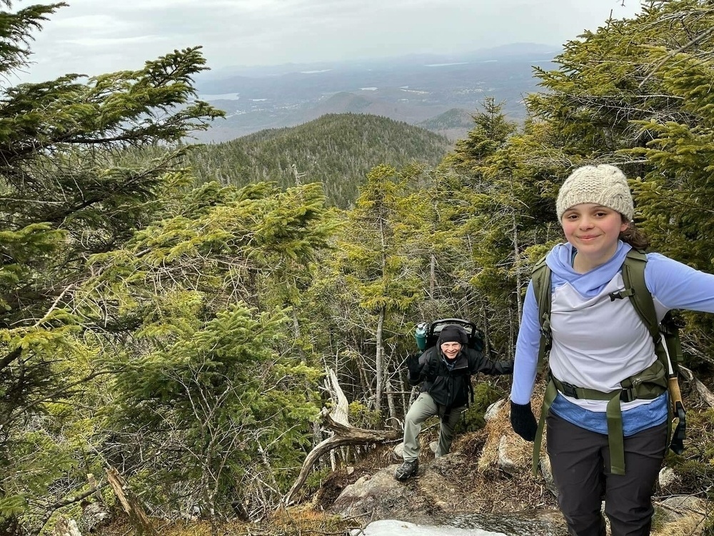 Two hikers ascending a forested mountain trail, with one in the foreground smiling at the camera, and expansive views of distant hills and a lake in the background.