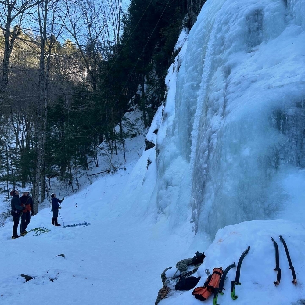 Three individuals stand near a tall ice formation in a snowy, wooded area, with ice climbing equipment laid out in the foreground.