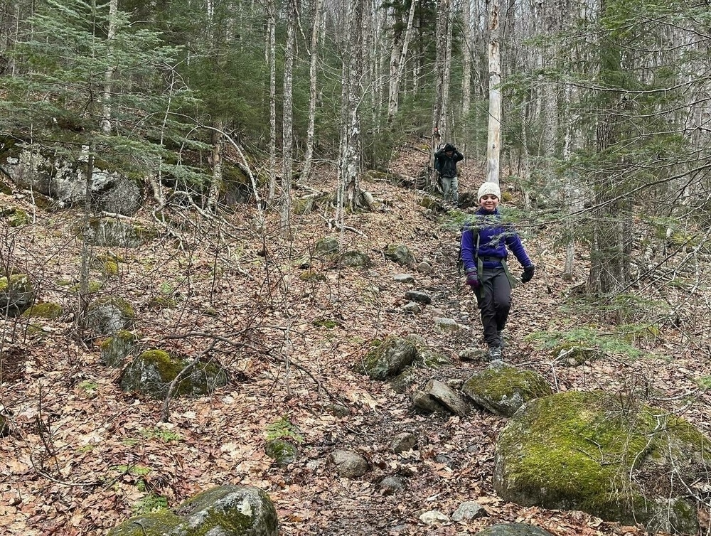 Two hikers are trekking on a rocky forest trail with bare trees and moss-covered rocks, one in the foreground smiling and one farther back.