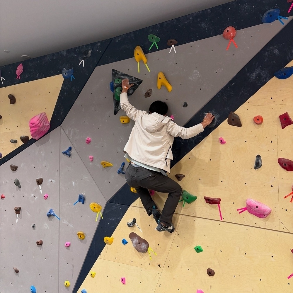 A person climbs a bouldering wall, gripping colorful handholds, in an indoor climbing gym.