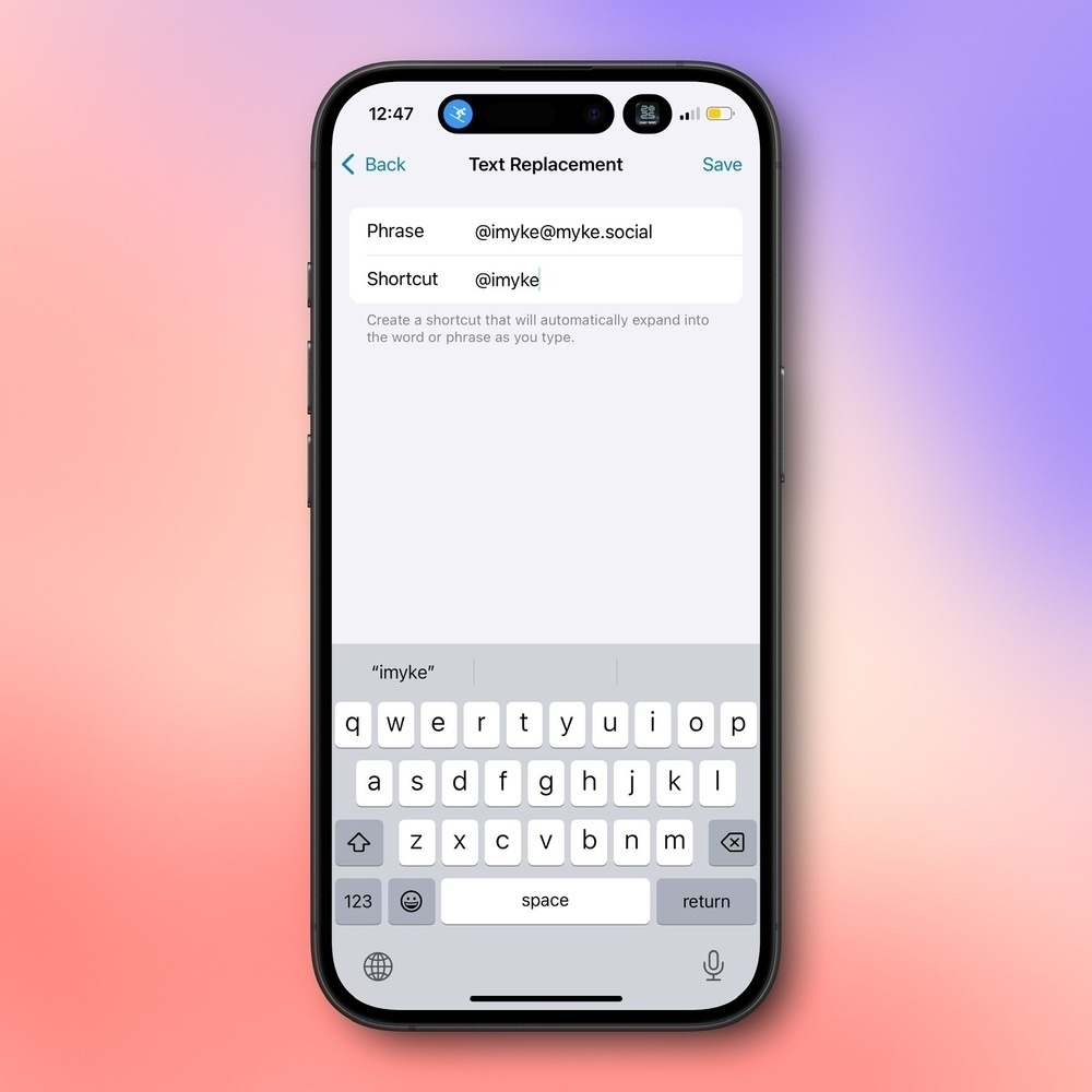 A smartphone displays a text replacement screen. The phrase &ldquo;@imyke@myke.social&rdquo; is set to expand from the shortcut &ldquo;@imyke&rdquo;. The backdrop is a gradient of purple and orange.