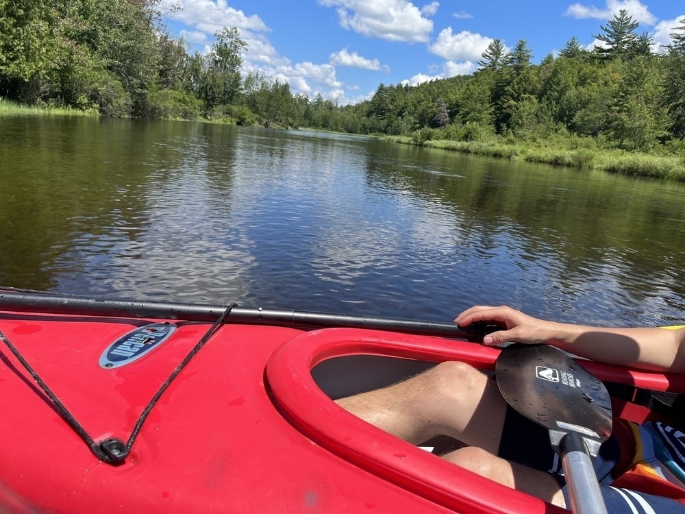 Person kayaking on a calm river, with lush green trees lining the banks under a blue sky with clouds.