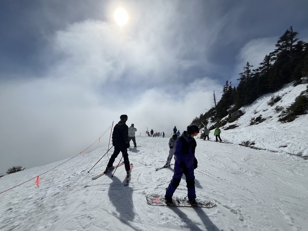 Skiers and snowboarders descend a snowy slope, with a bright sun above and dense clouds parting in a mountainous setting.