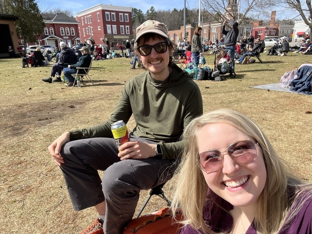 Two people are smiling for a selfie in a sunny park with other people relaxing on the grass, against a backdrop of buildings and trees.