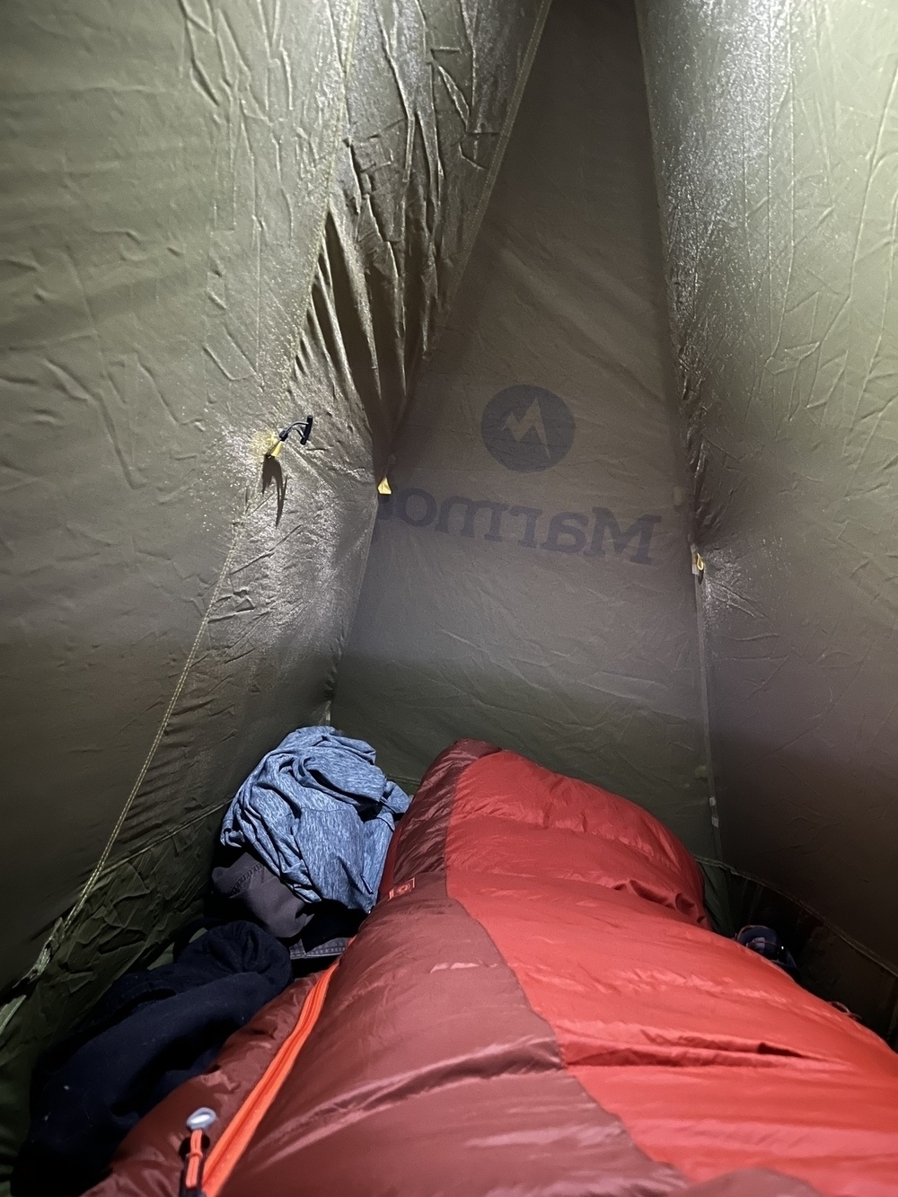 Inside a tent, a person views their sleeping space with a red sleeping bag, clothing, and the tent&rsquo;s fabric inner walls featuring a logo shadow.