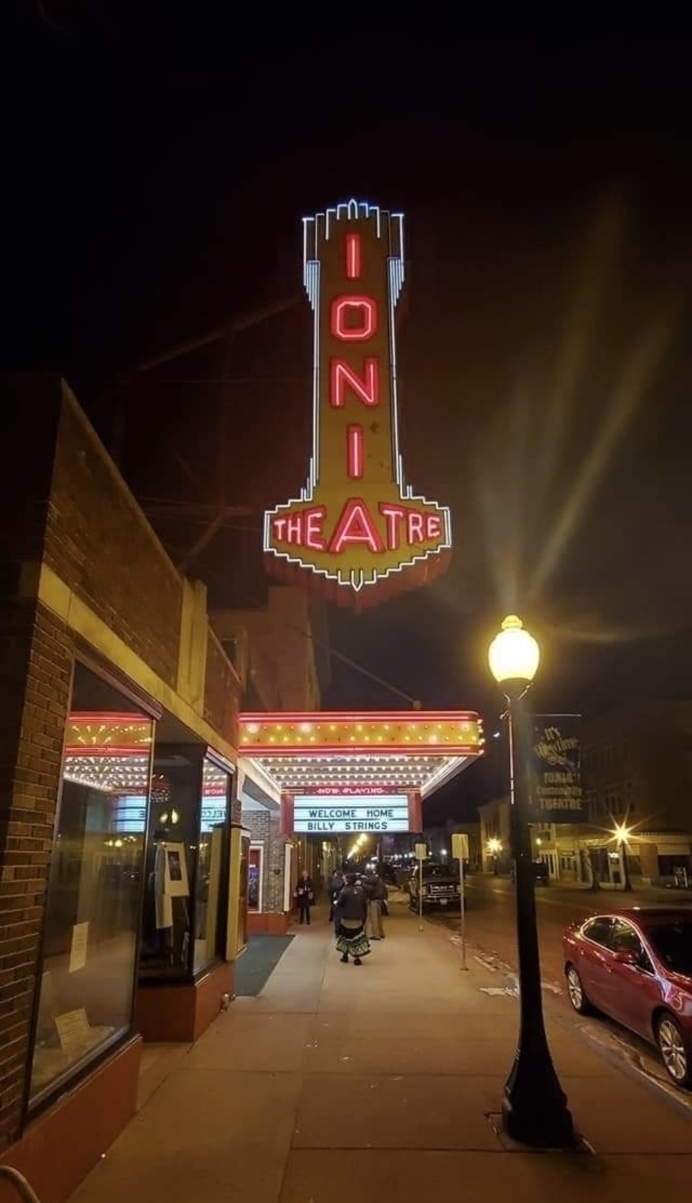 Neon-lit theater marquee at night reads &ldquo;IONIA THEATRE&rdquo; with &ldquo;WELCOME HOME BILLY STRINGS&rdquo; on a lower sign, flanked by street lights and a pedestrian sidewalk.