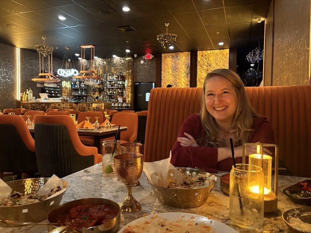 A smiling woman seated at a dining table with plates of food in a dimly lit restaurant with elegant decor.