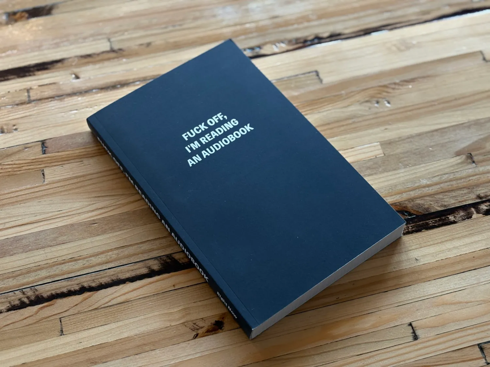 A black book lies closed on a wooden table. White text on the cover reads, &ldquo;FUCK OFF, I&rsquo;M READING AN AUDIOBOOK.&quot;