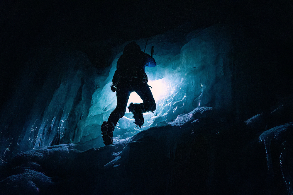 A figure in climbing gear is mid-climb up a dark, blue-tinged ice column.