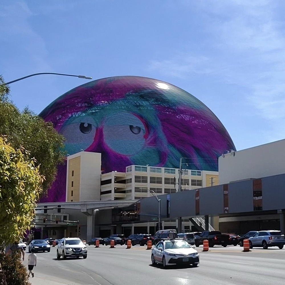 A large whimsical building screen on the Sphere in Las Vegas depicts eyes, set against a clear sky with active city streets and vehicles below.