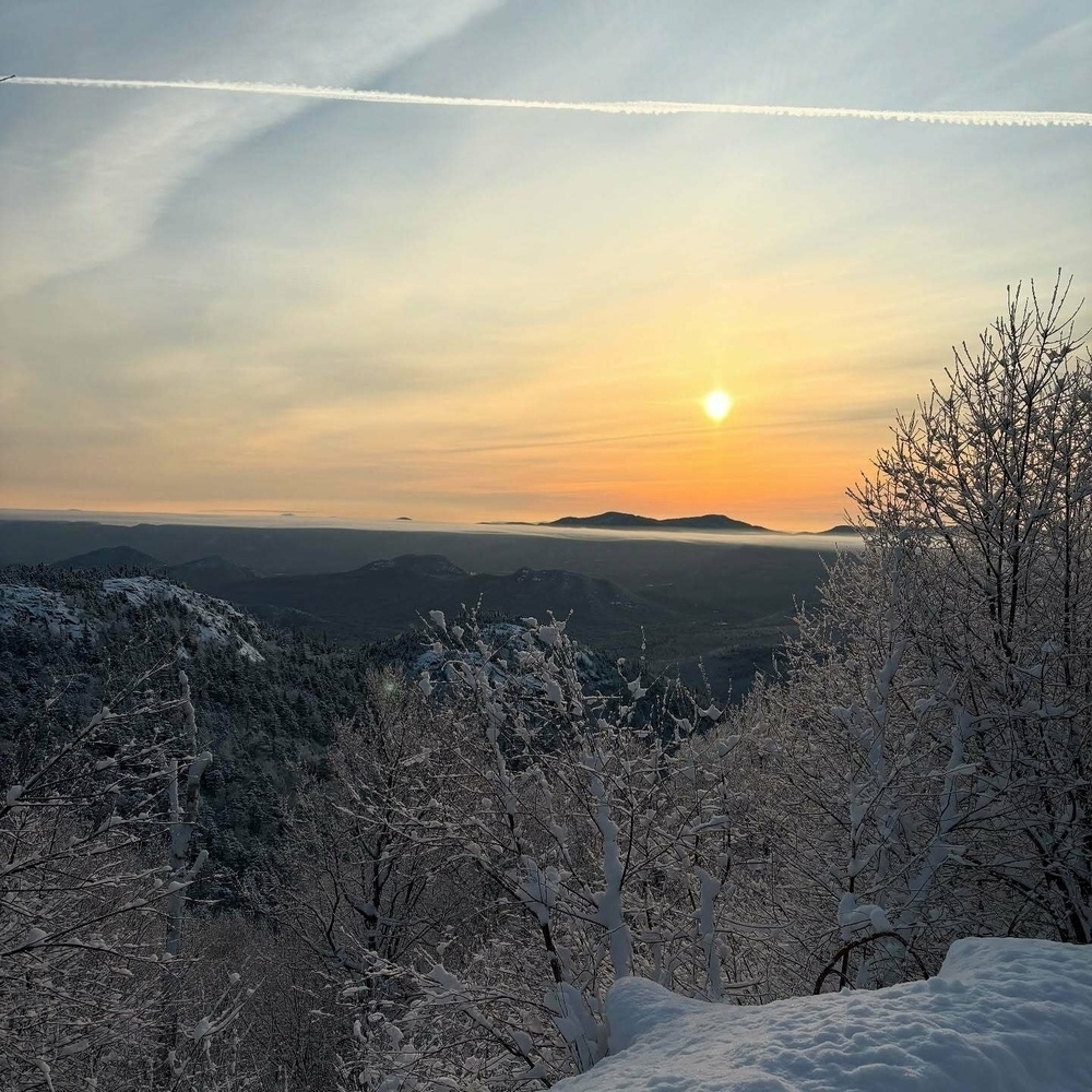 Sun setting over snow-covered trees with distant mountains in the background under a clear sky. A vapor trail stretches across the sky, creating a peaceful winter scene.