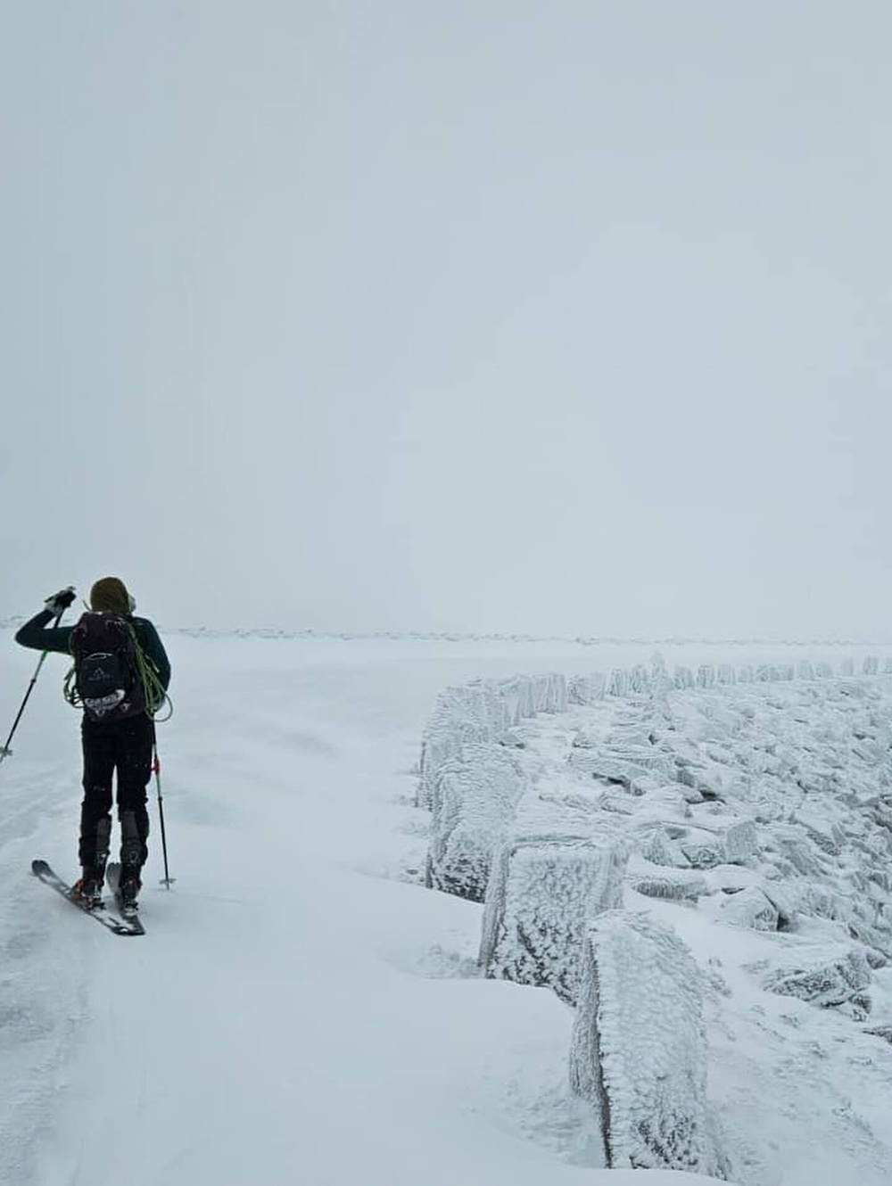 A skier gliding across a snow-covered landscape with a backpack traverses a path beside a frost-covered rocky embankment under an overcast sky.