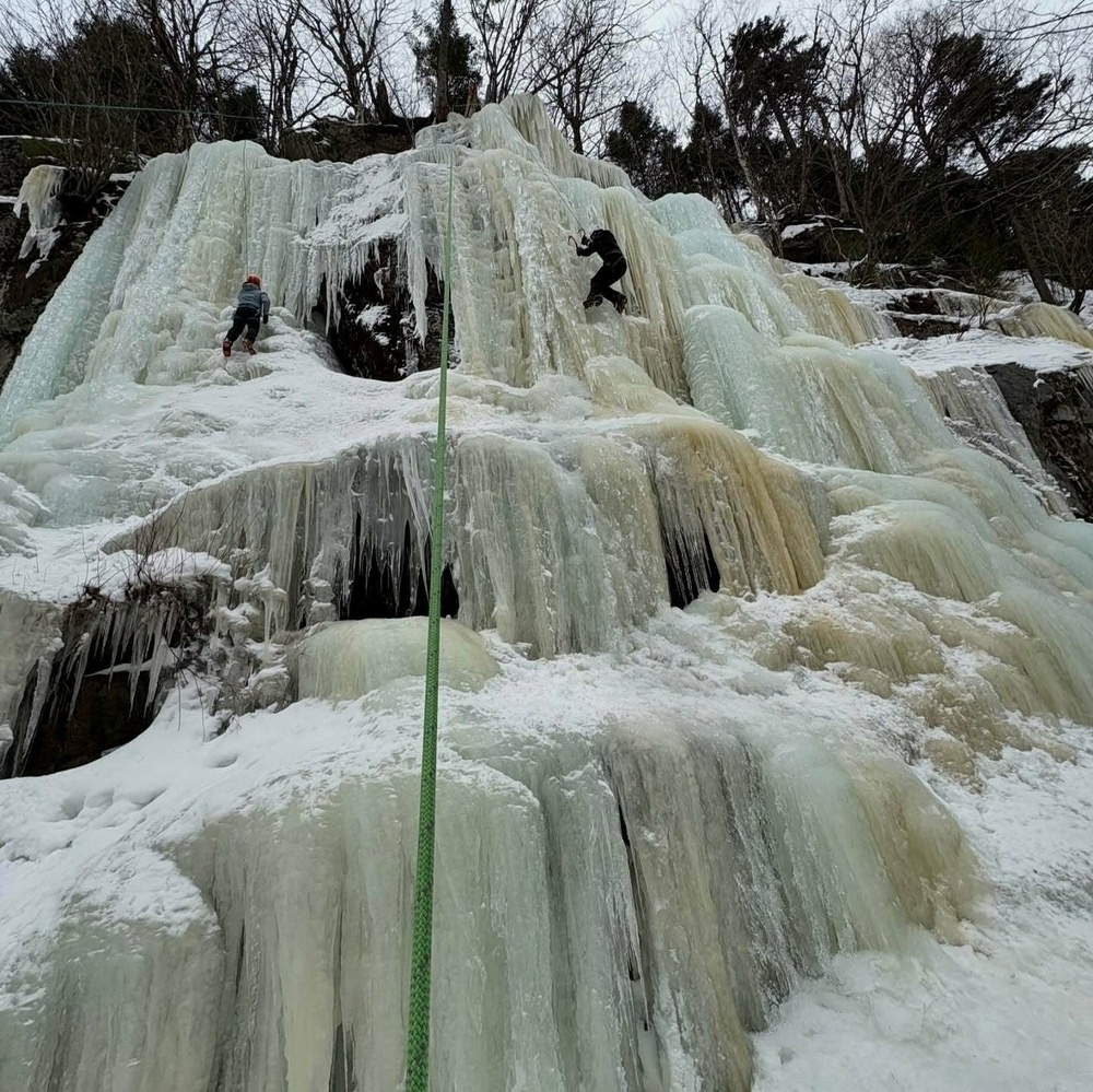 Two people are ice climbing on a vertical frozen waterfall using ropes amid a snow-covered landscape with trees above.
